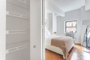 Ein Schlafzimmer mit einem Bett, einem Fenster mit Blick auf Gebäude und einer Tür zu einem Schrank mit Regalen.