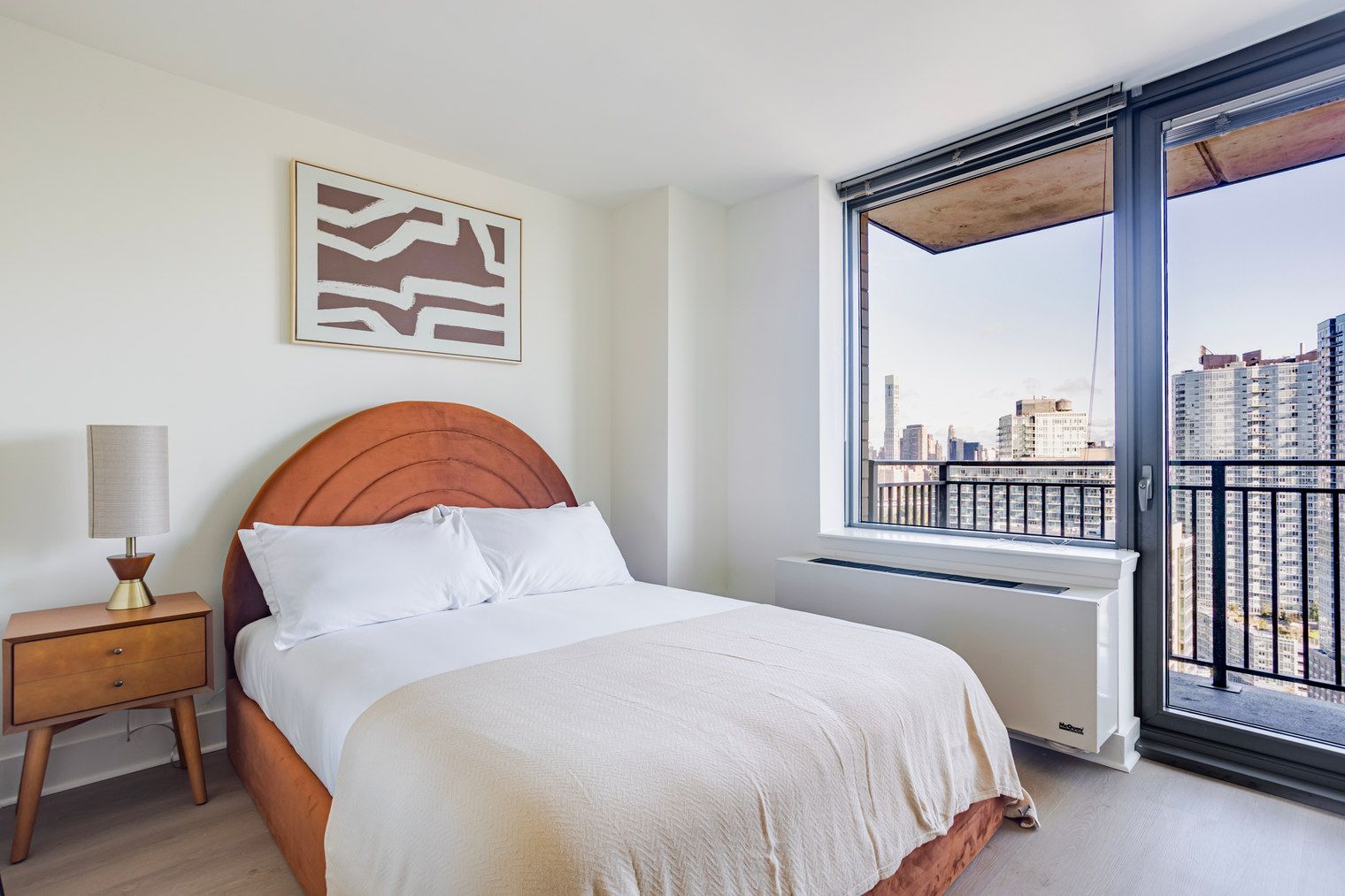 A bedroom with a bed, a nightstand, a lamp, a window with a view of buildings, and a radiator.