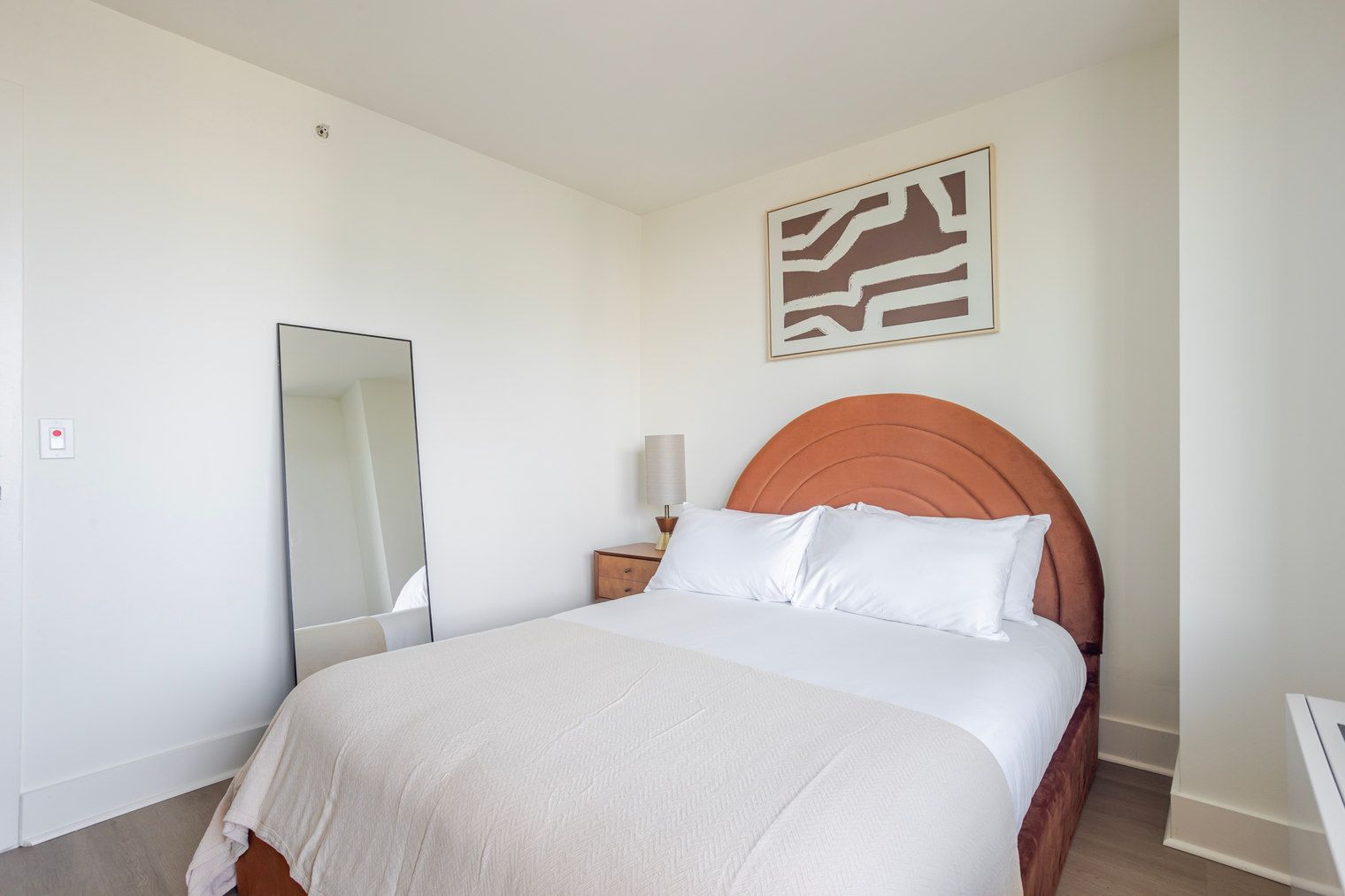 A bedroom with a bed, a nightstand, a mirror, and a wall-mounted artwork.