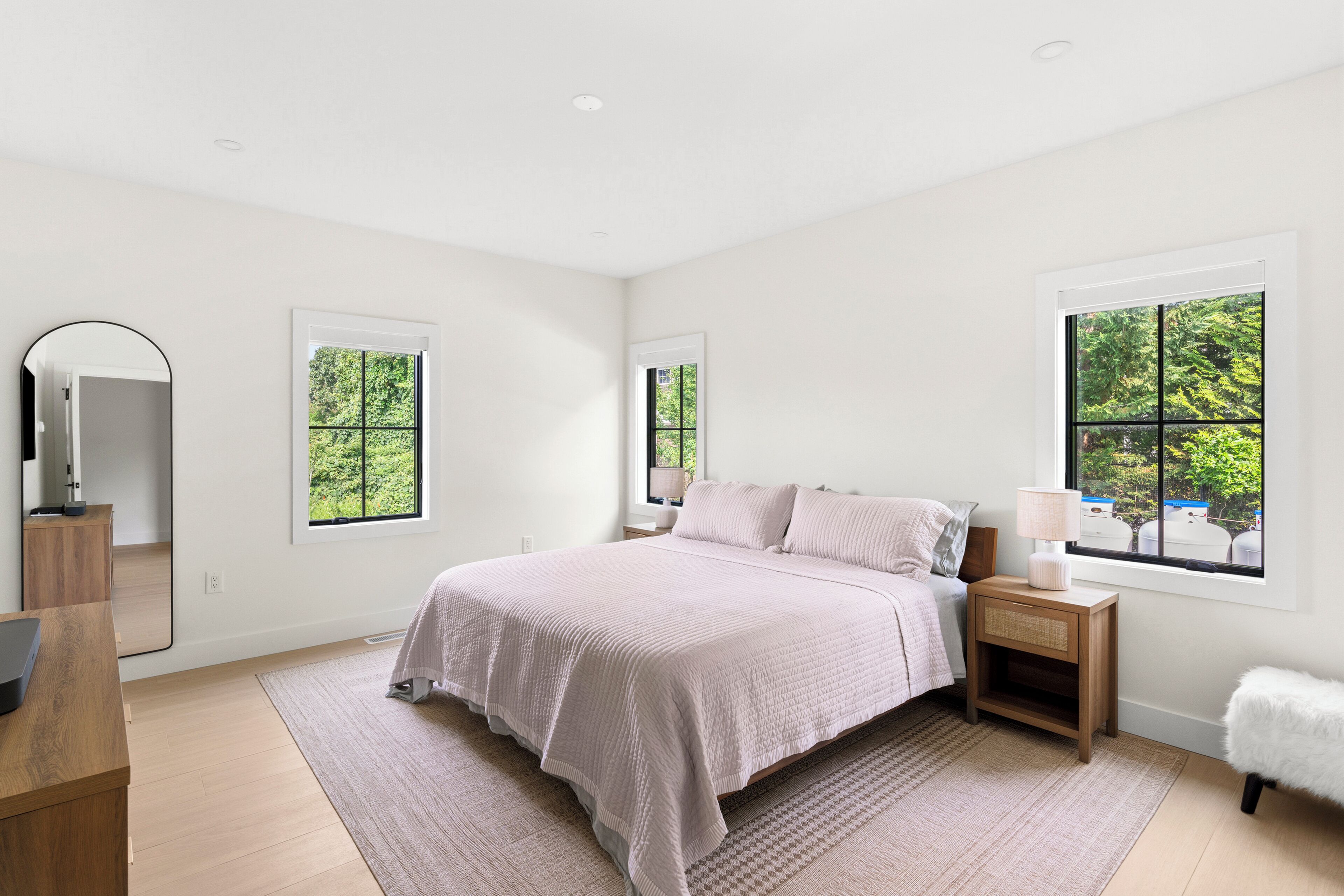 Une chambre moderne avec un grand lit, des tables de chevet, un miroir et des fenêtres donnant sur un paysage verdoyant.