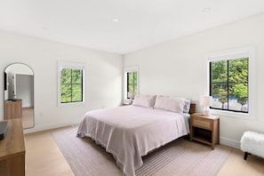 A modern bedroom with a large bed, bedside tables, a mirror, and windows with a view of greenery.