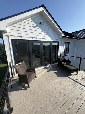 A covered deck with two wicker chairs and a bench, a sliding glass door, and a white exterior wall.