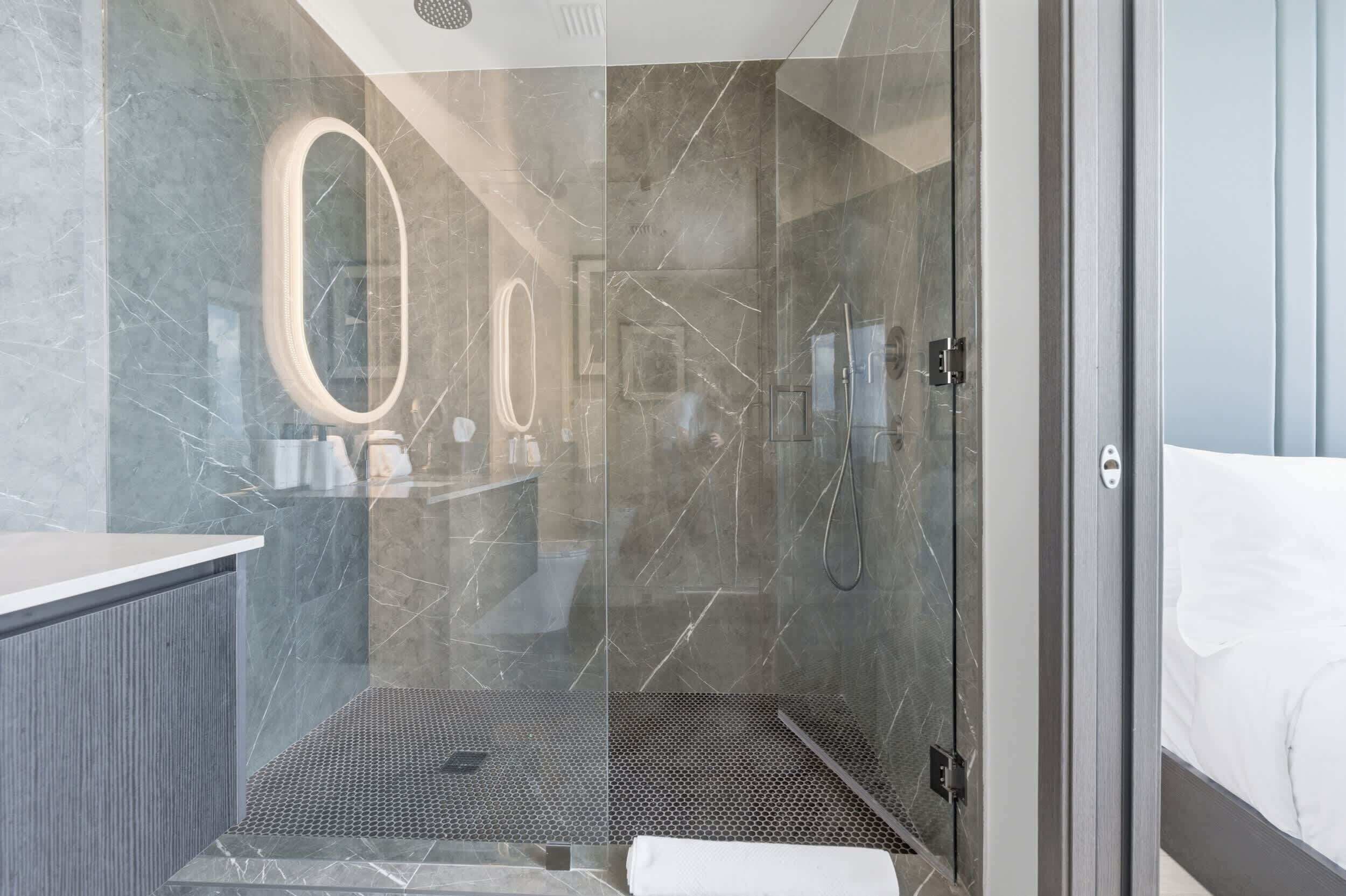 A modern bathroom with a glass-enclosed shower, a marble-tiled wall, and a large mirror.