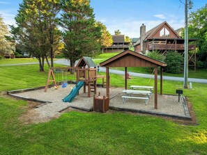 Une aire de jeux avec un toboggan et des balançoires, entourée d’une pelouse et d’arbres.