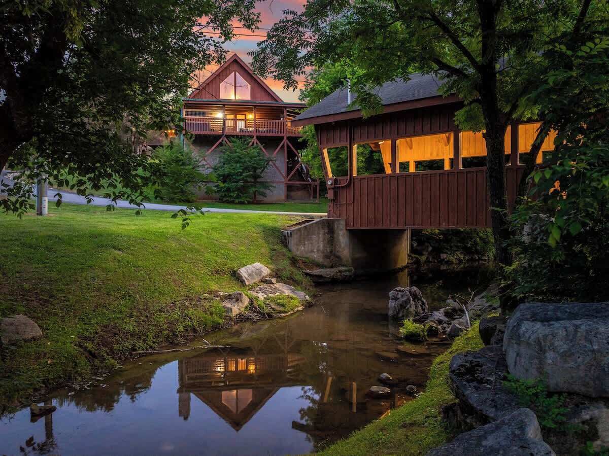 A wooden bridge over a calm stream, a grassy area, and a building with a sloped roof in the background.