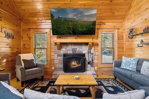 A cozy cabin interior with a stone fireplace, a flat-screen TV mounted above it, and a comfortable seating area with a sofa and armchairs. - Creekside Villa Retreat with Hot Tub, Fire Pit and Playground (Sevierville)