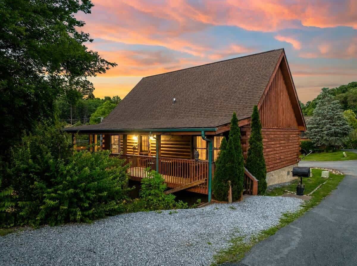 A log cabin with a porch, surrounded by greenery and a gravel driveway.