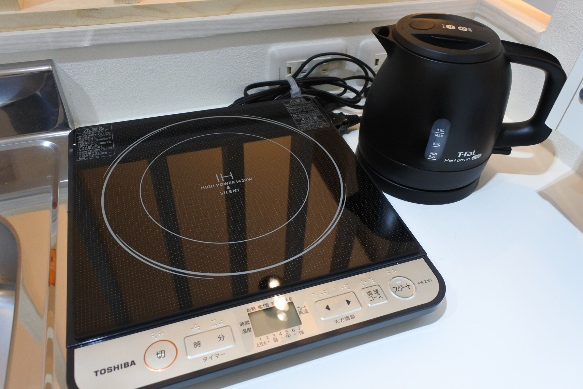 Eine Toshiba digitale Küchenwaage mit schwarzem und silbernem Design, auf einer weißen Arbeitsplatte neben einem schwarzen Wasserkocher.