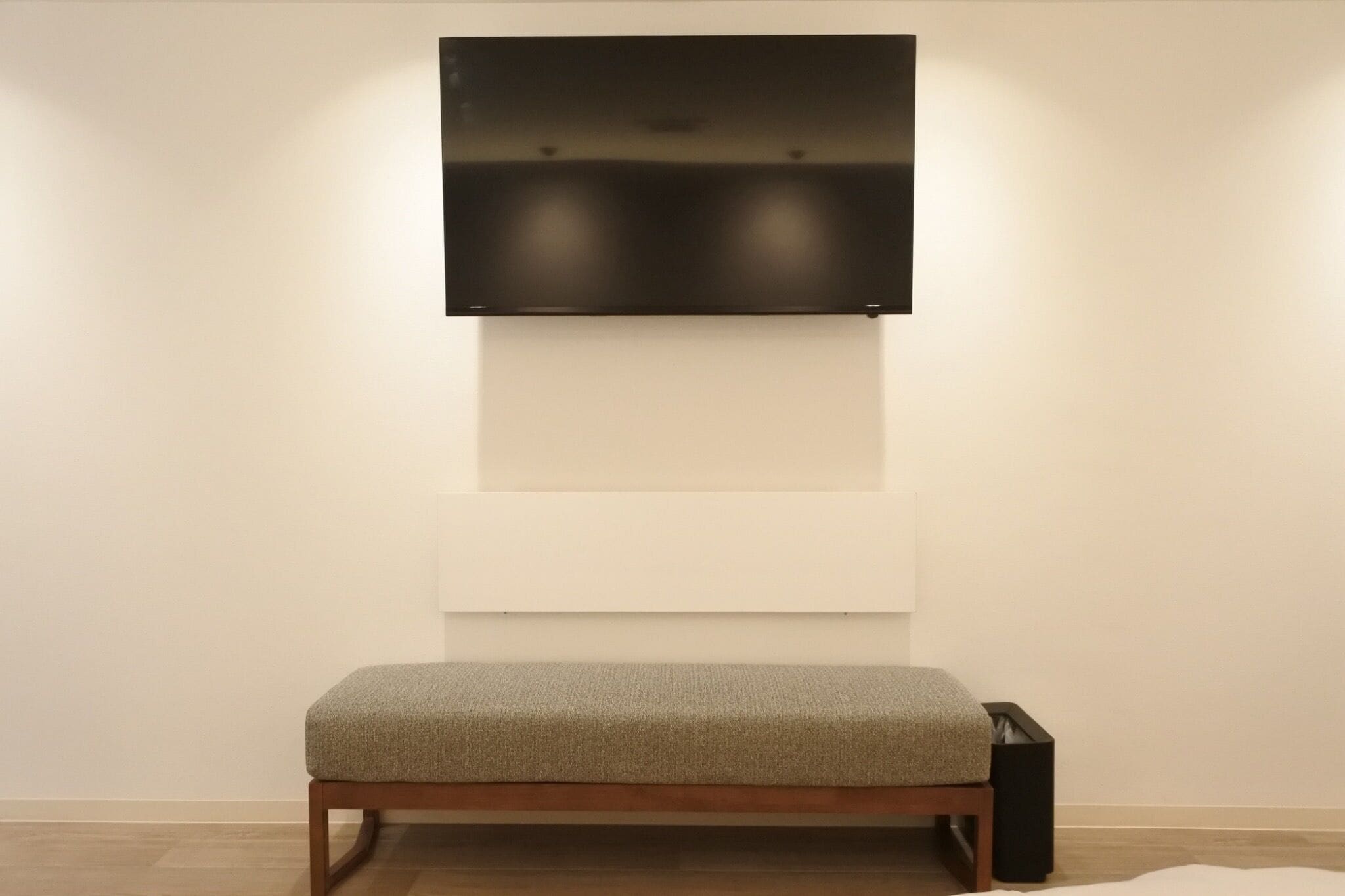A minimalist room with a bench, a flat-screen TV mounted on the wall, and a trash bin.