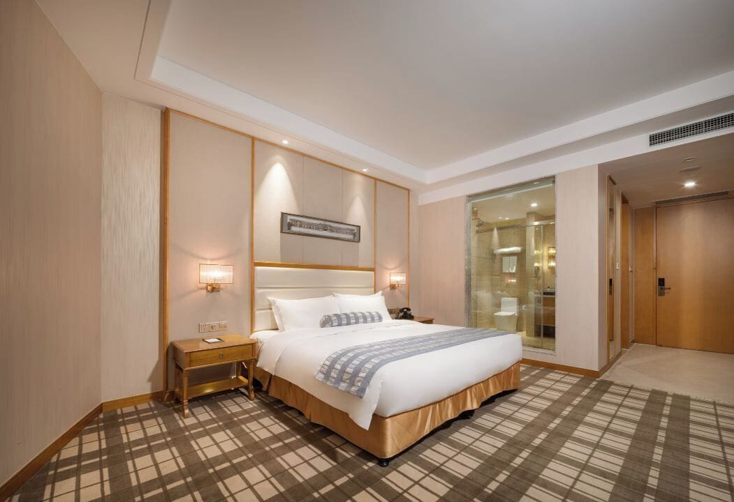 Une chambre d’hôtel avec un grand lit, des tables de chevet, une petite table, une lampe fixée au mur, un tableau encadré et une cloison en verre.