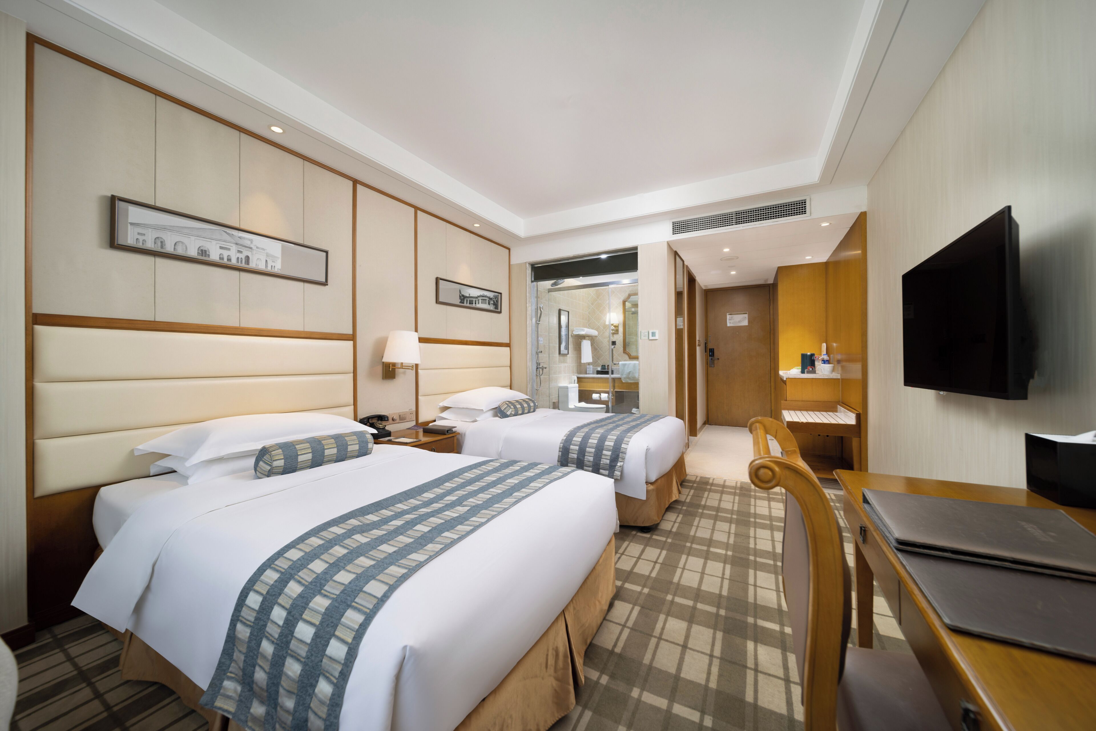 Quarto de hotel com duas camas, uma escrivaninha, uma cadeira, uma televisão e um banheiro visível através de uma porta aberta.