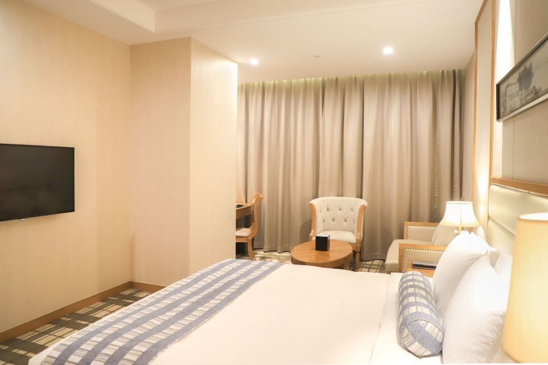 Habitación de hotel con cama, televisión, silla, mesa y ventana con cortinas.
