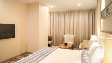 Habitación de hotel con cama, televisión, silla, mesa y ventana con cortinas.
