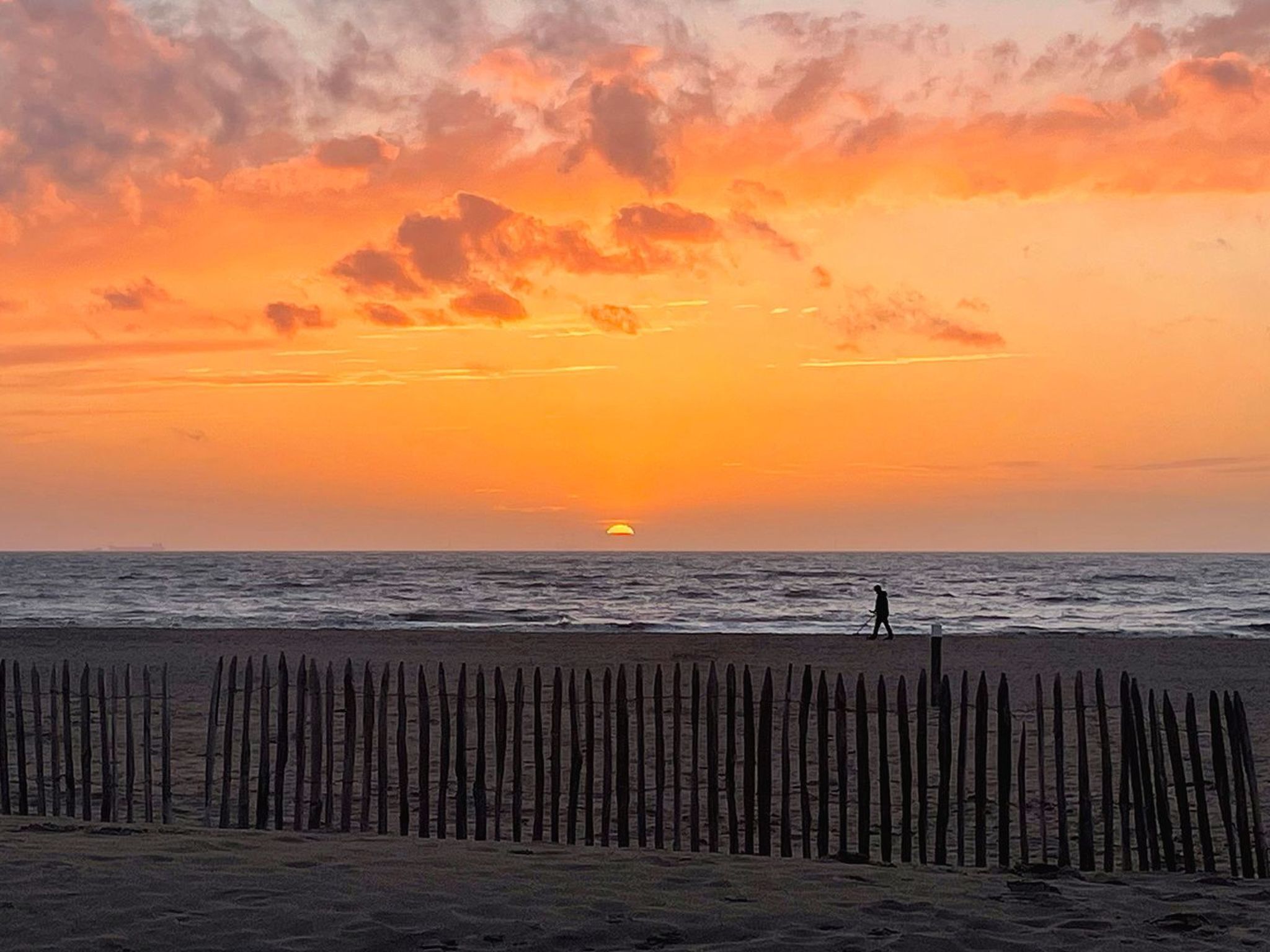 A sunset over the ocean with a person walking on the beach and a wooden fence in the foreground.