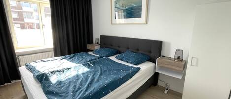 Un dormitorio con cama, mesita de noche, lámpara, un cuadro enmarcado en la pared y una ventana con cortinas.