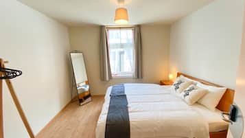Ein Schlafzimmer mit einem Bett, einem Spiegel, einem Nachttisch, einer Lampe und einem Fenster mit Vorhängen.