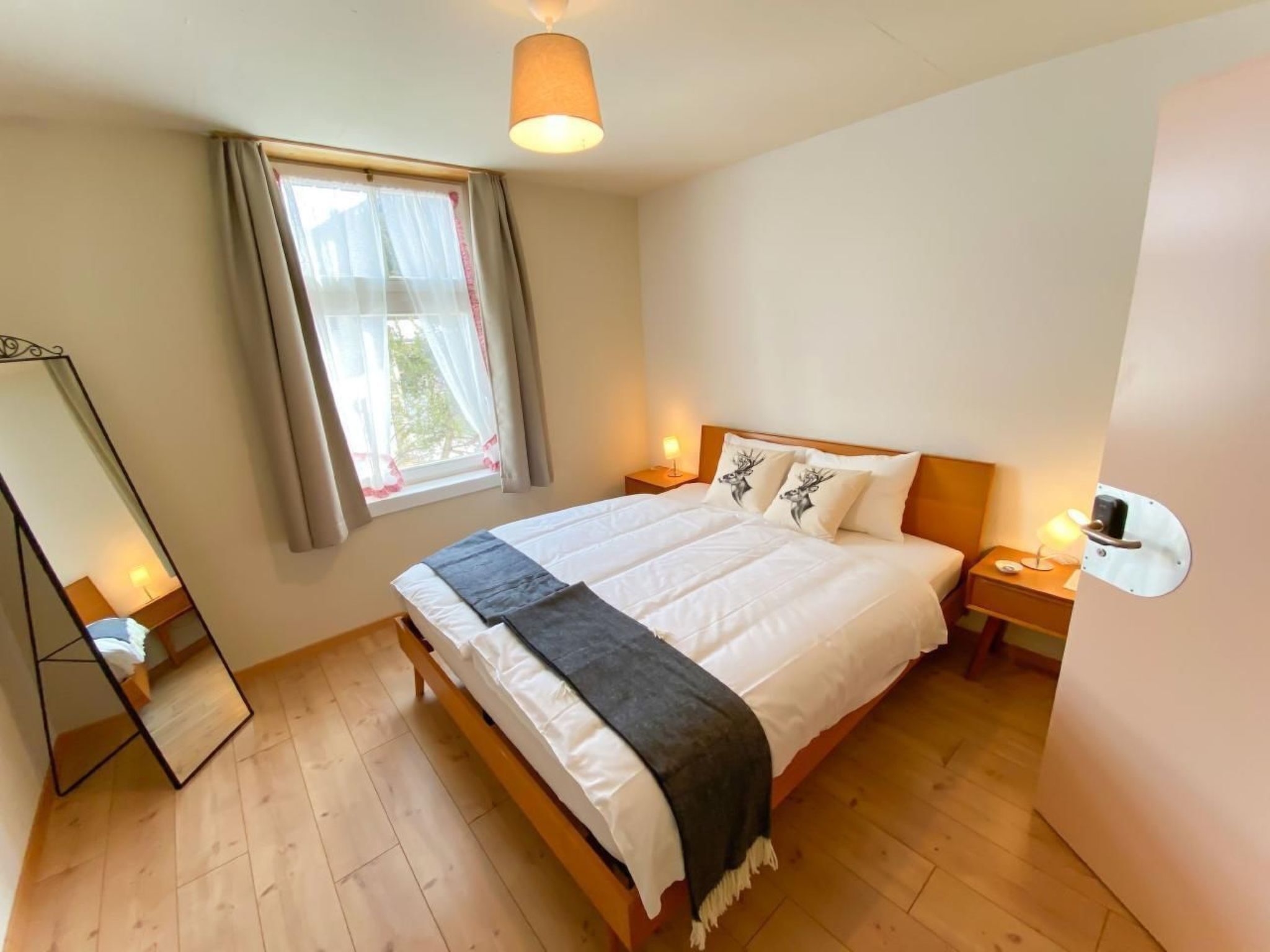 Ein Schlafzimmer mit einem Bett, Nachttischlampen, einem Fenster mit Vorhängen, einem Spiegel und einem Holzboden.