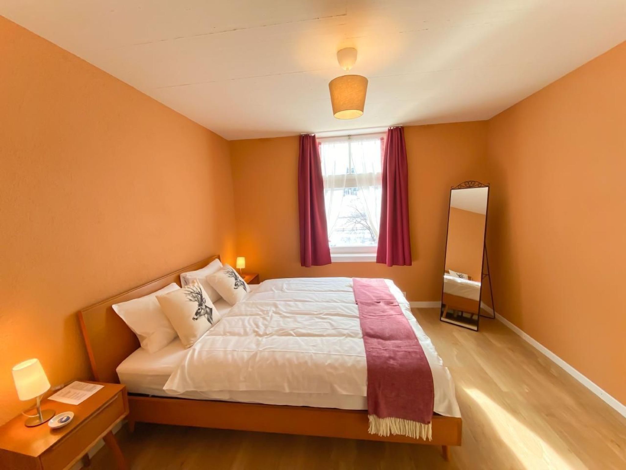 Ein Schlafzimmer mit einem Bett, einem Nachttisch, einer Lampe, einem Fenster mit Vorhängen und einem Spiegel.