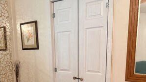 Quarto com porta dupla, quadro emoldurado na parede, um vaso com uma cruz e piso de madeira.