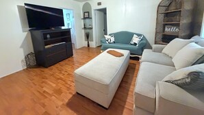 Uma sala de estar com televisão de tela plana, sofá, um banquinho e um ventilador de teto.