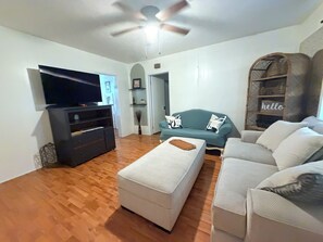 Ein Wohnzimmer mit Flachbildfernseher, einer Couch, einem Hocker und einem Deckenventilator.