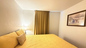 Quarto de hotel com cama, luminária, quadro e janela com cortinas.