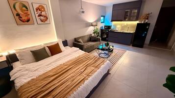 A modern bedroom with a large bed, a sofa, a small coffee table, and a kitchen area in the background.