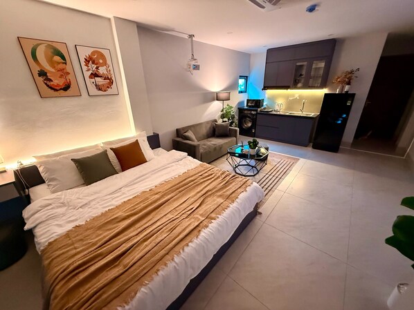 A modern bedroom with a large bed, a sofa, a small coffee table, and a kitchen area in the background.