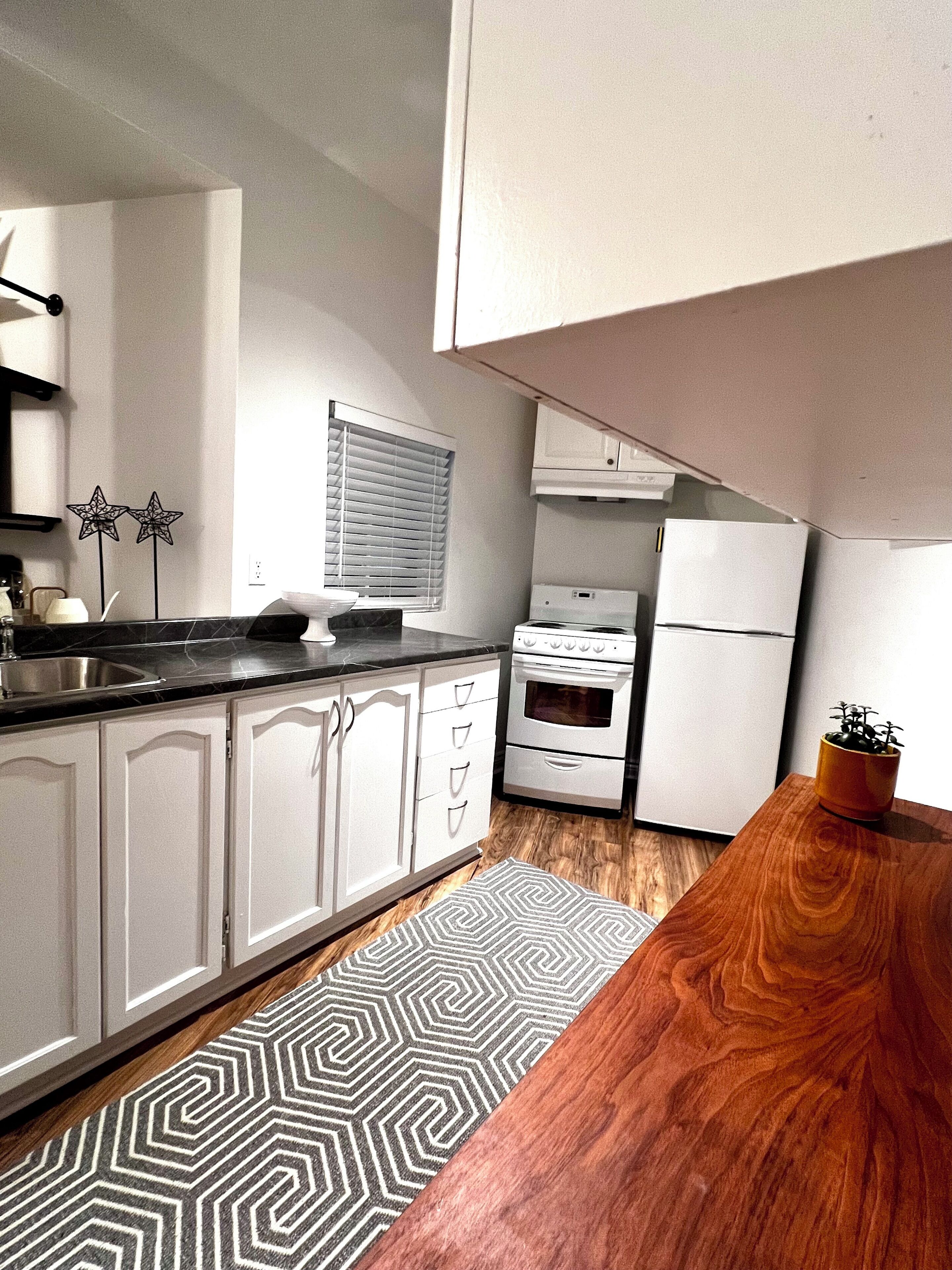 A kitchen with white cabinets, a black countertop, a stove, a refrigerator, and a window with blinds.