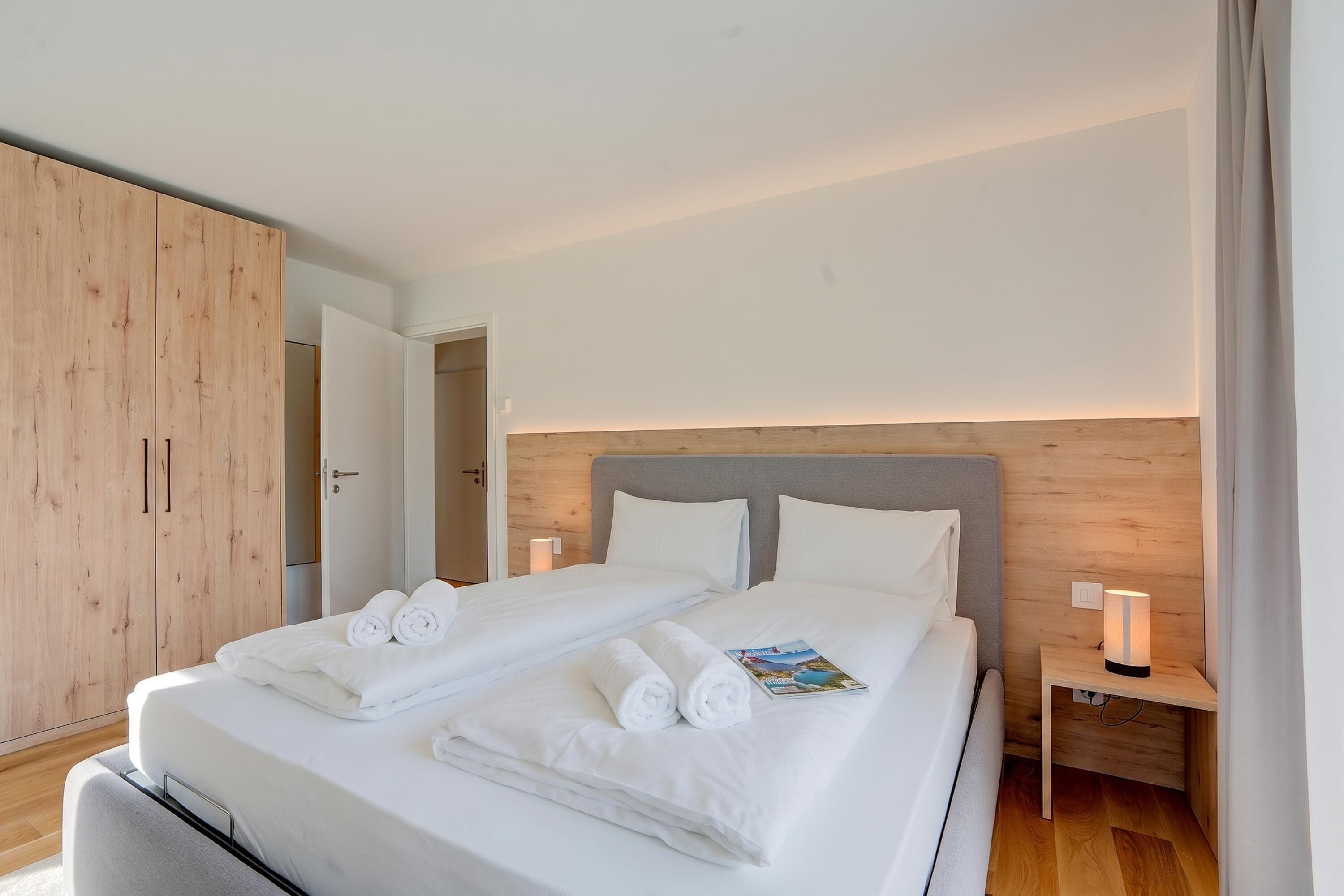 A double bed with white linens and towels, a wooden wardrobe, a bedside table with a lamp, and a nightstand.