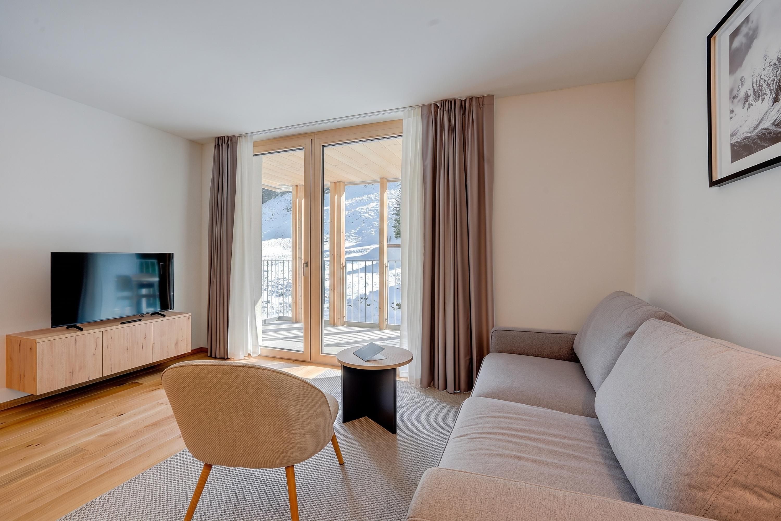 Un salon moderne doté d’un téléviseur à écran plat, d’un canapé, d’un fauteuil et offrant une vue sur un paysage enneigé grâce à de grandes portes vitrées.
