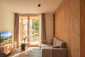 Ein modernes Wohnzimmer mit Holzboden, einer Couch, einem Fernseher und Blick auf einen Balkon mit Holzwertkasten.
