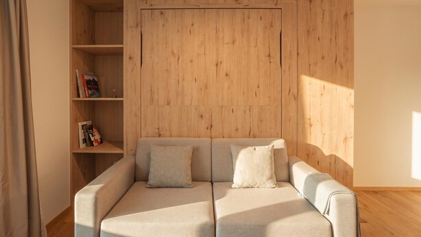 A modern living room with a sofa, a wooden bookshelf, and a large window. - Residenza Castanea 208 Modern Alpine Stay in San Bernardino | Ski, Hiking & Nature, San Bernardino, Switzerland (San Bernardino)