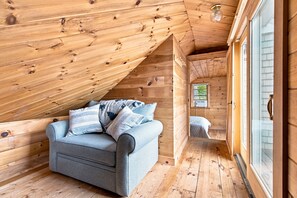 A cozy attic bedroom with a wooden ceiling, a gray sofa with pillows, and a window with blinds. - Rare 4-bed, 3-full bath Cottage & Tiny Home w/ Private Beach & Dock (Damariscotta)