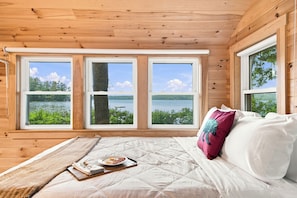A wooden cabin room with a bed, a breakfast tray, and three windows overlooking a lake.