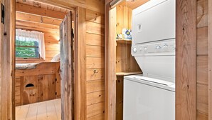 Interior de una cabaña de madera con una cama, una ventana con cortina y un electrodoméstico blanco.