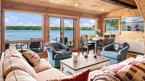 Una cabaña con interior de madera, ventanas amplias, zona de comedor y vistas a un lago.