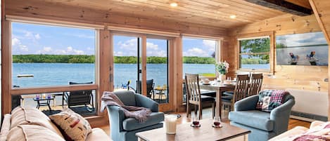 Une cabane avec un intérieur en bois, de grandes fenêtres, un coin repas et une vue sur un lac.