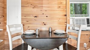 Interior de una cabaña de madera con una mesa de comedor, sillas y una ventana que da a un paisaje de árboles.