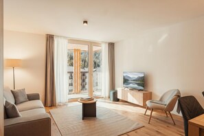 Ein modernes Wohnzimmer mit einer Couch, einem Couchtisch, einem Fernseher und Blick auf einen Wald durch große Fenster.