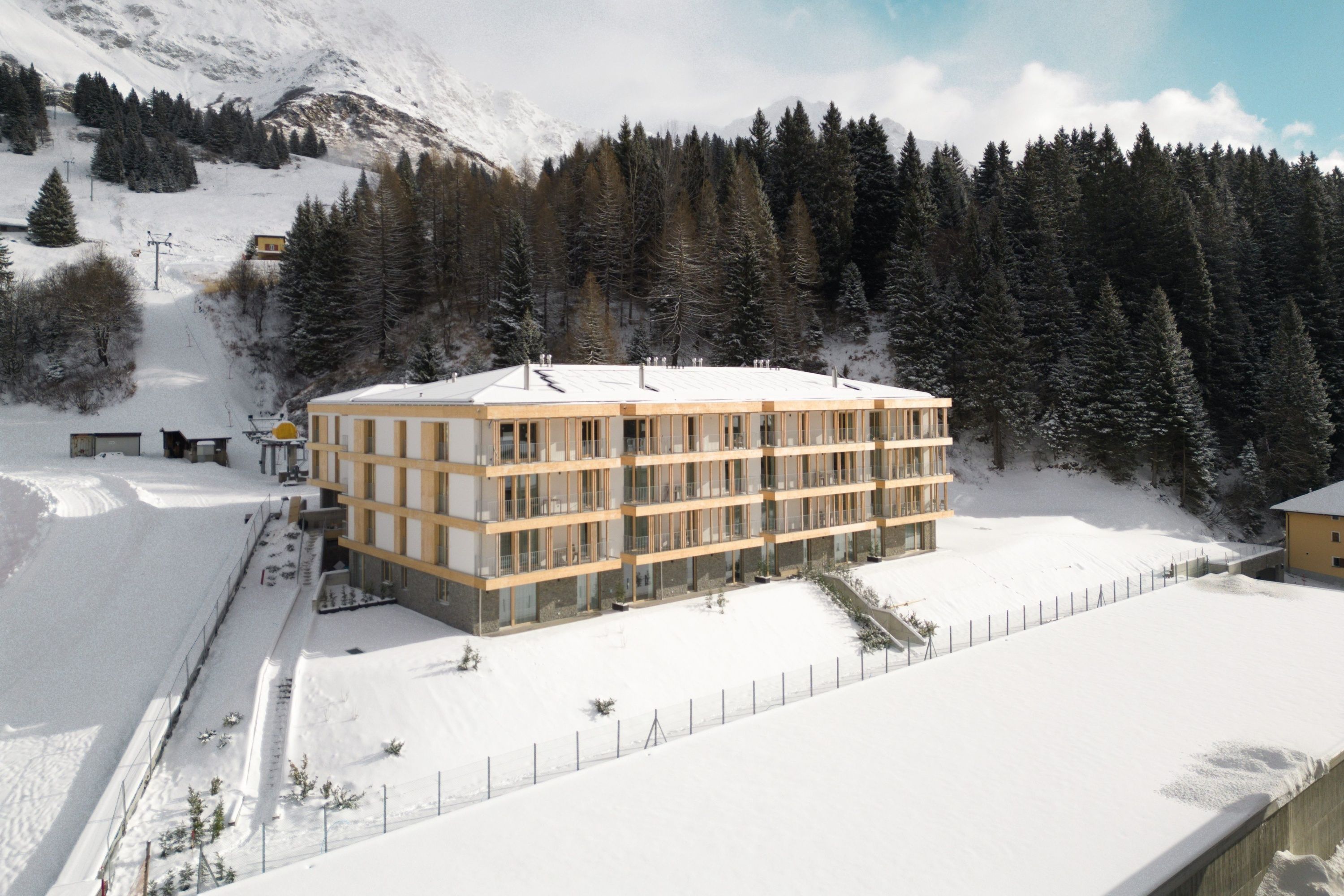 A multi-story hotel with a flat roof, surrounded by snow-covered ground and a forested mountain backdrop.