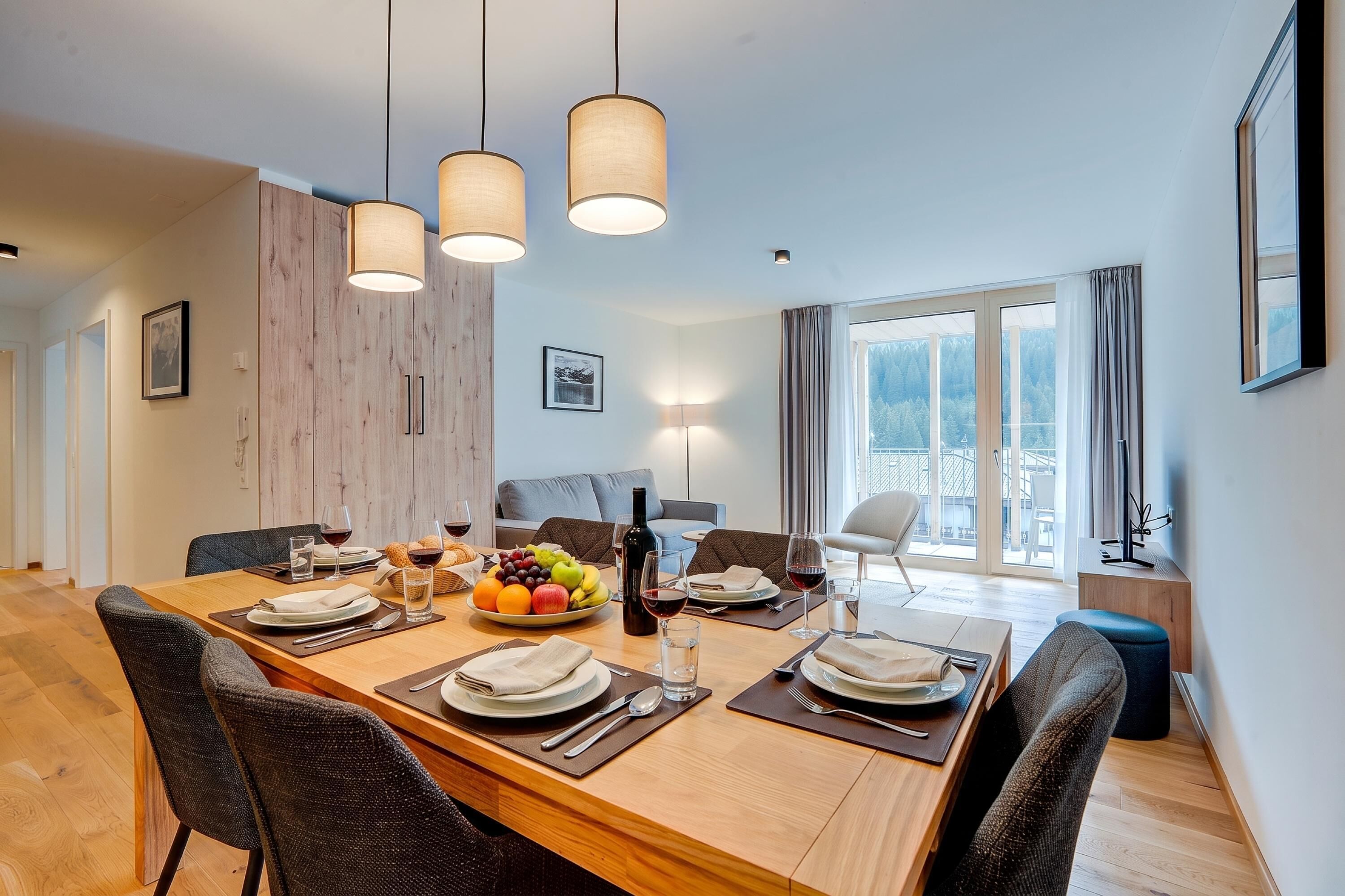 A dining area with a wooden table set for four, surrounded by chairs, with a view of a forest through large windows.
