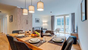 A dining area with a wooden table set for four, surrounded by chairs, with a view of a forest through large windows.