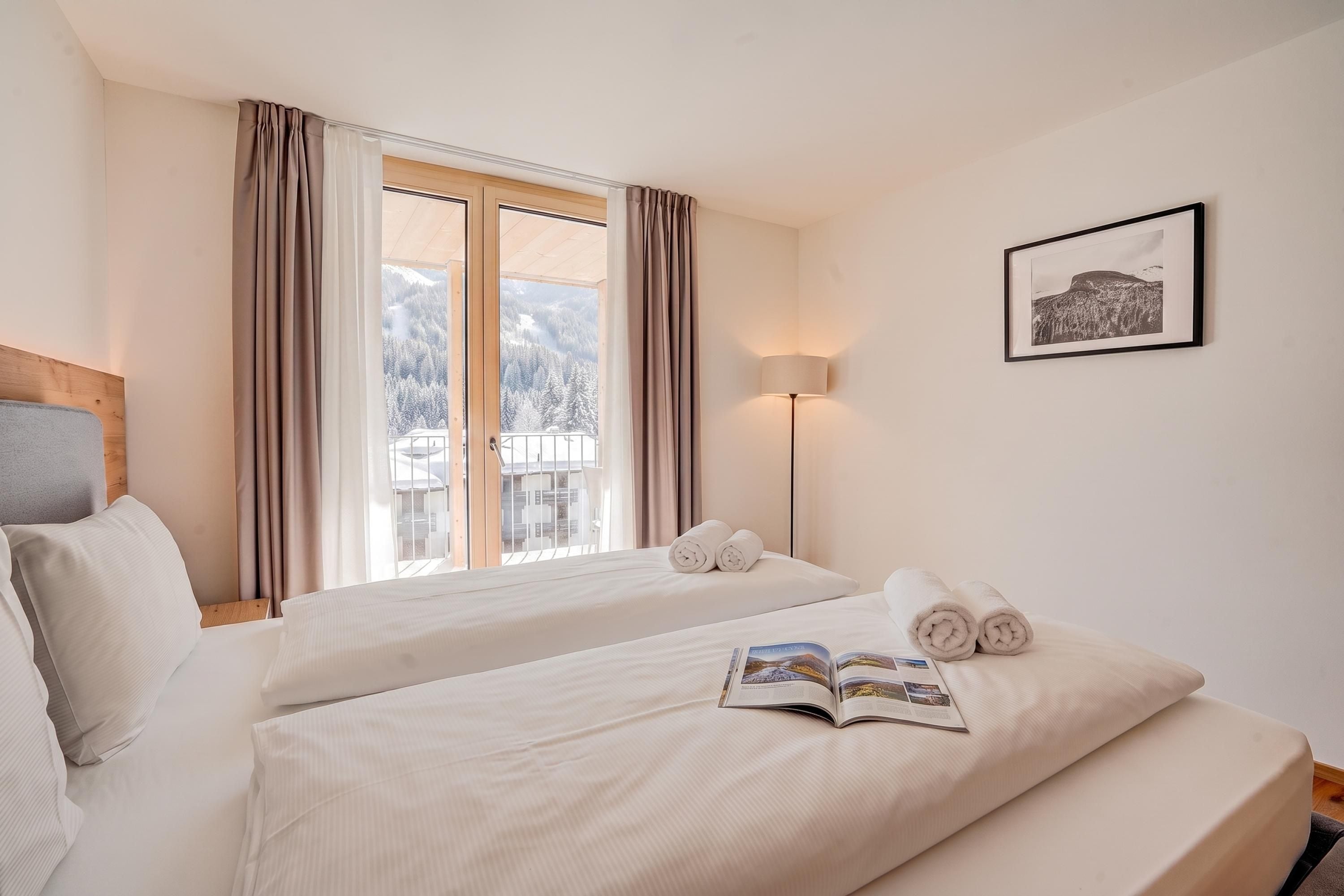 A hotel room with two beds, a window with curtains, a bedside lamp, a framed picture on the wall, and a magazine on one of the beds.
