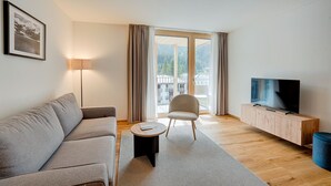 Een moderne woonkamer met een bank, een klein rond tafeltje, een stoel, een televisie op een houten kast en een groot raam met gordijnen.