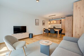 Ein modernes Wohnzimmer mit Flachbildfernseher, grauen Sofas, einem Couchtisch und einem Essbereich mit Holztisch und Stühlen.