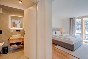 A modern hotel room with a large bed, a bathroom with a sink and mirror, and a view of a snowy landscape through the window. - Residenza Castanea 202 San Bernardino Alpine Apartments, San Bernardino, Switzerland (San Bernardino)