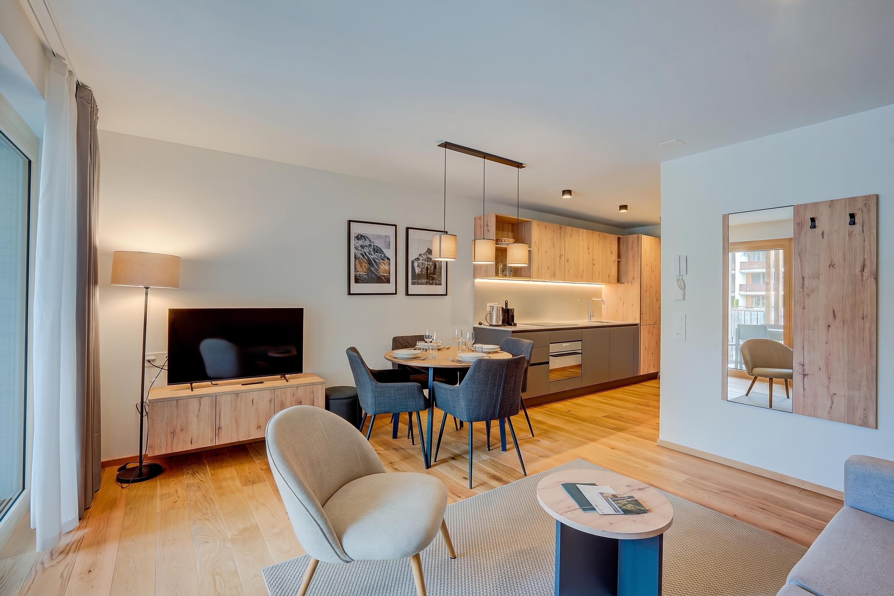 Un salon moderne doté d’un téléviseur à écran plat, d’un coin repas avec une table et des chaises, et d’une cuisine avec des meubles en bois et un éclairage suspendu.
