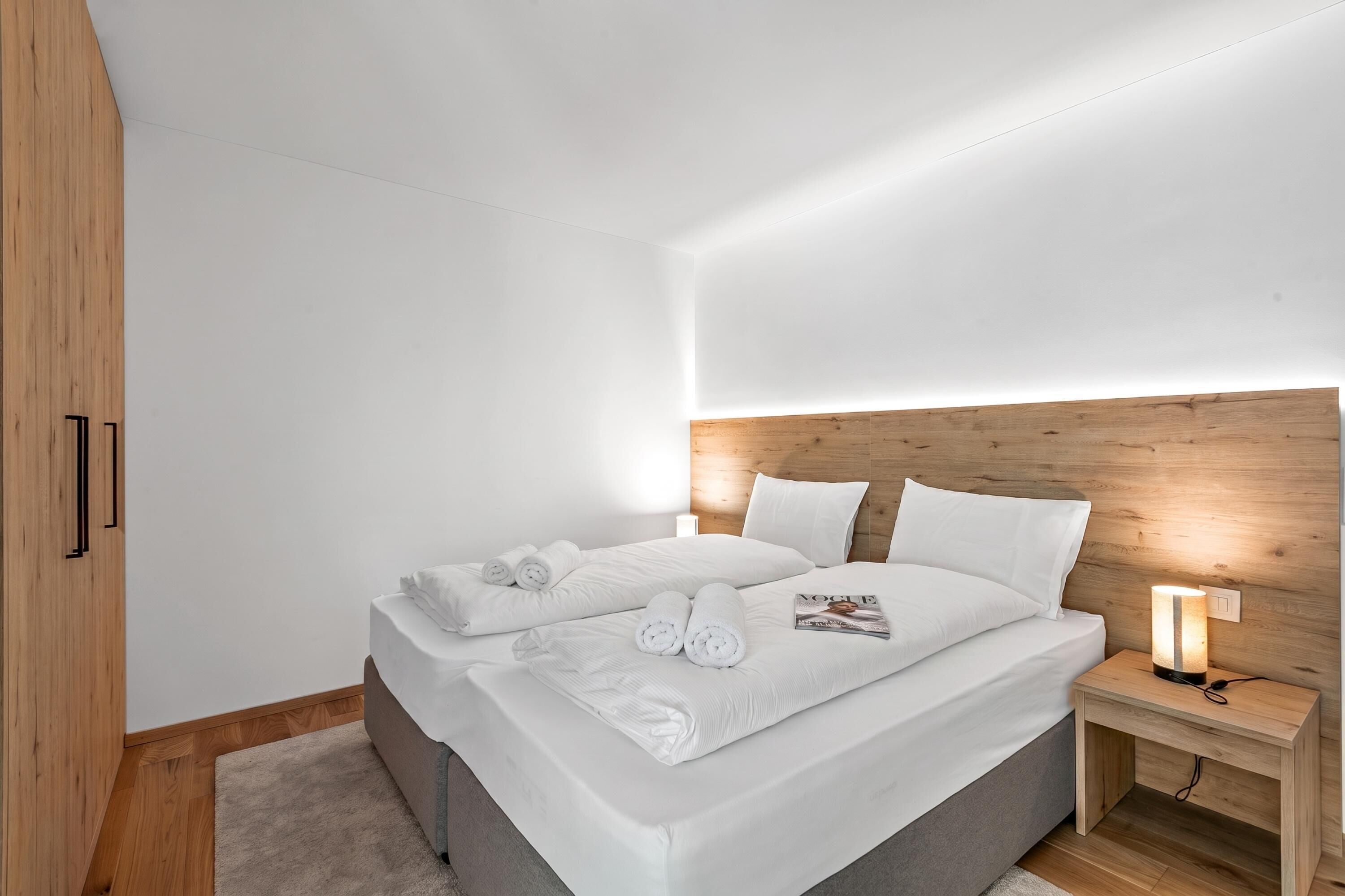 A modern bedroom with a wooden headboard, a bed with white linens, and a bedside table with a lamp.
