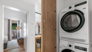 A laundry room with a front-loading washer and dryer, a wooden door, and a view into a living area with a sofa, table, and chair. - Residenza Castanea 107 Modern Alpine Stay in San Bernardino | Ski, Hiking & Nature, San Bernardino, Switzerland (San Bernardino)